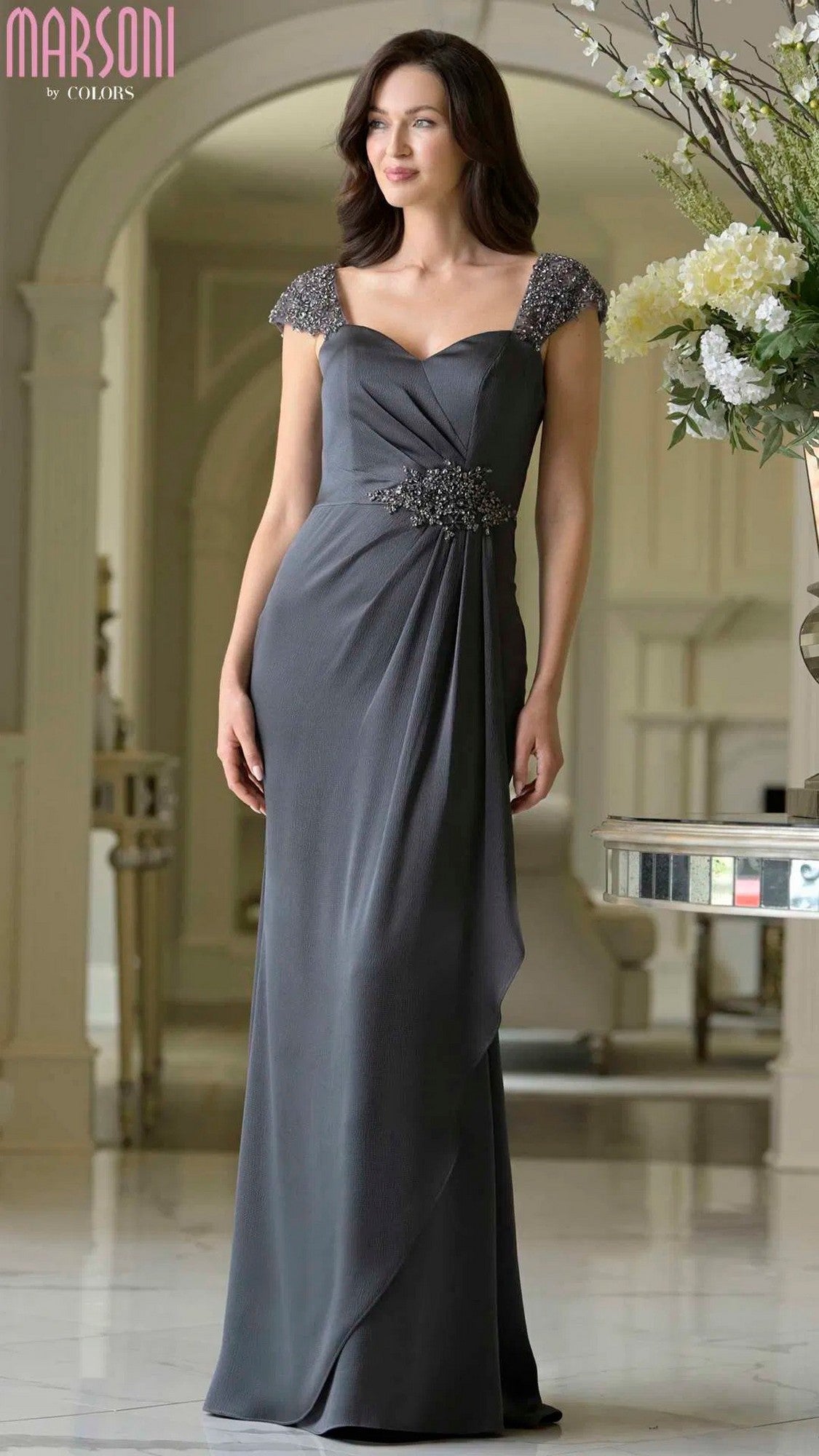 A woman stands indoors wearing the Marsoni MV1377, a charcoal long formal dress with an empire waist, beaded cap sleeves, and decorative waist detail.
